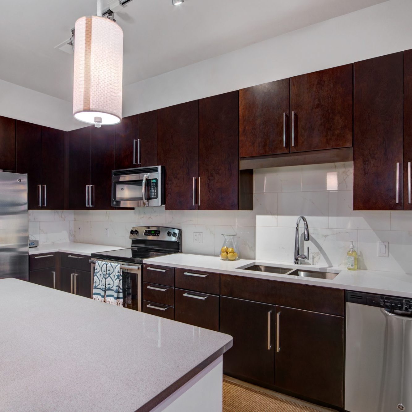 Dark wood cabinets accent this modern home kitchen remodel. Stainless steel appliances and fixtures with light colored counter tops and tile splash back.